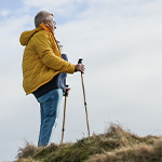 Senior hiker standing on a grassy hill with trekking poles, wearing a yellow jacket and looking into the distance.